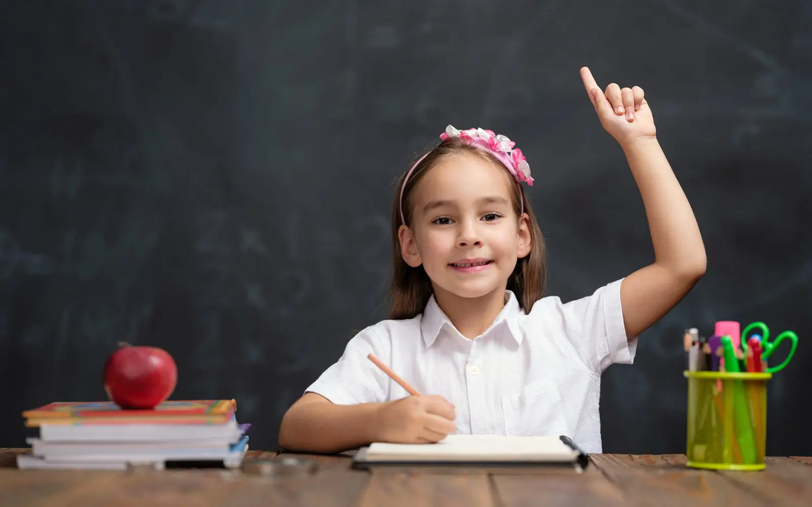 Child raising hand in classroom, smiling confidently.
